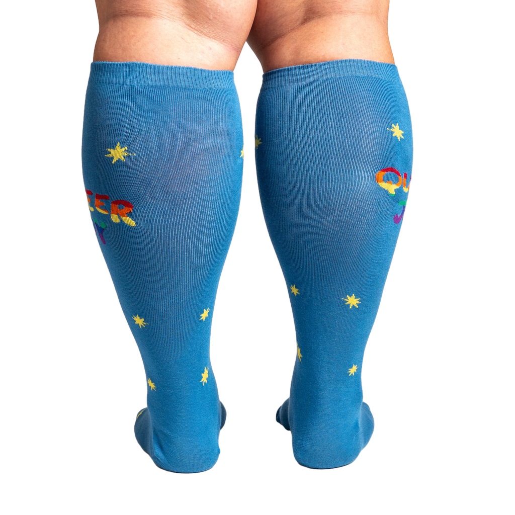 Rear view of model wearing blue knee-high socks featuring 'Queer Joy' in rainbow and yellow stars.