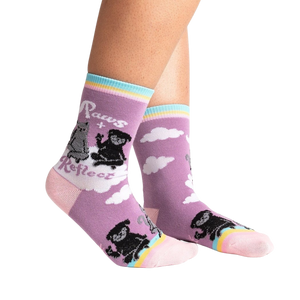 A pair of purple socks with a black dog and a gray cat on them. The socks have a rainbow and clouds at the top and a pink toe and heel.