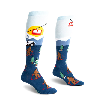 front view of a pair of knee-high socks. the socks have a blue background with brown bigfoot figures skiing down a snowy mountain. there are also small green trees and a red ski lift in the image. the top of the sock is light blue.