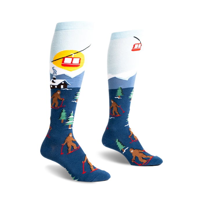 front view of a pair of knee-high socks. the socks have a blue background with brown bigfoot figures skiing down a snowy mountain. there are also small green trees and a red ski lift in the image. the top of the sock is light blue.