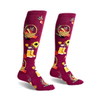 Pair of burgundy knee-high socks with autumn harvest design: pumpkins, sunflowers, baskets, and yellow boots.