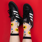 [lf] Feet in black Adidas Sambas and sheer sunflower and bee socks against a red background.