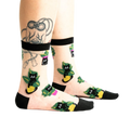 A pair of sheer ankle socks with a black band at the top, featuring a whimsical repeating pattern of black cats in yellow and green plant pots.