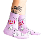A person's tattooed legs wearing the light lavender "Hey Boo!" ghost crew socks. A "Glows in the Dark" star graphic is visible.