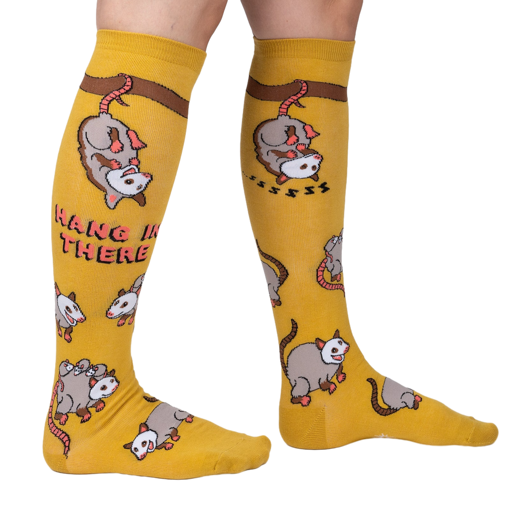Lifestyle shot: Model wearing a pair of mustard yellow knee-high socks with possum designs.