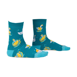 Pair of teal crew socks with a whimsical pattern of geese floating on inner tubes and swimming. Yellow and white geese on teal background.