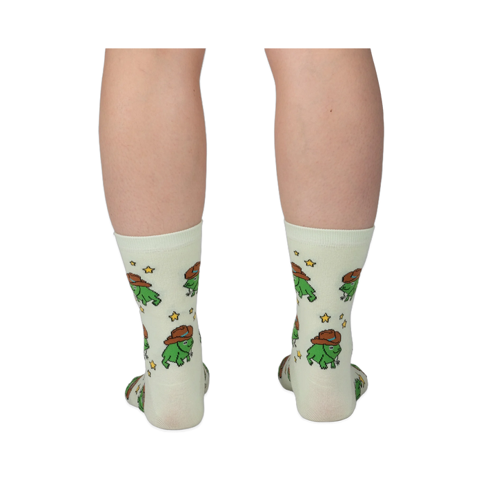 Back view of legs wearing pale crew socks featuring a pattern of green pixel frogs in brown cowboy hats and yellow stars.