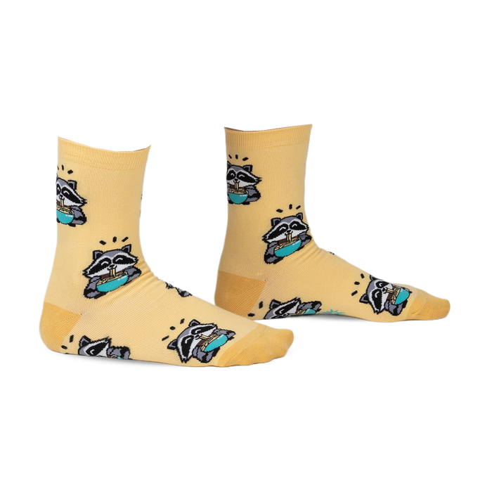 Pair of pastel yellow crew socks featuring a repeating pattern of raccoons enjoying ramen. 