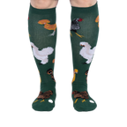 Person wearing dark green knee-high socks patterned with various chickens and eggs, standing, front view.