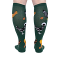 Person wearing dark green knee-high socks patterned with various chickens and eggs, standing, back view.