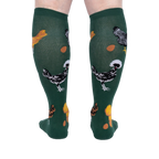 Person wearing dark green knee-high socks patterned with various chickens and eggs, standing, back view.