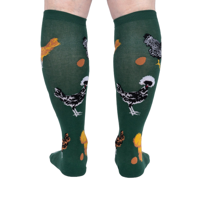 Person wearing dark green knee-high socks patterned with various chickens and eggs, standing, back view.