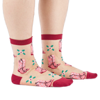 A person's legs and feet, wearing sheer pink cowboy boot socks from the side. Legs are bare against a white background.