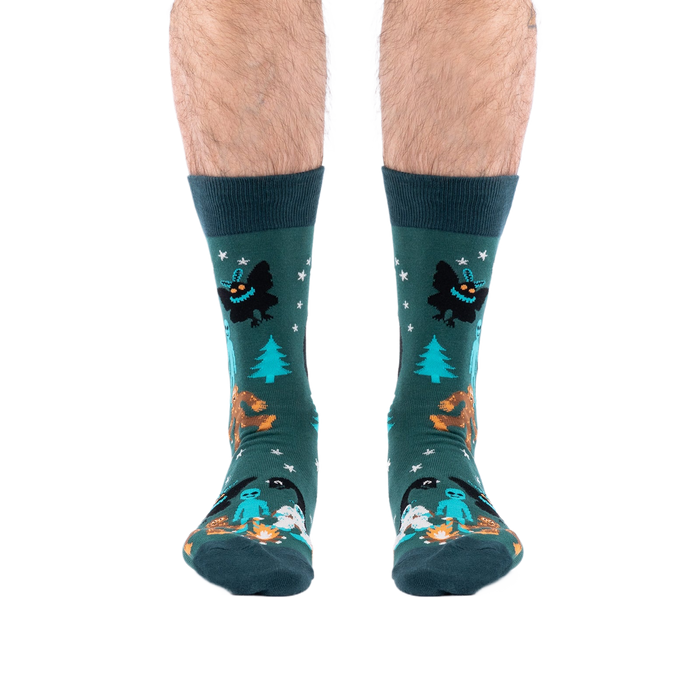 Front view of a person's lower legs, showing both dark green cryptid socks featuring Bigfoot and Mothman.
