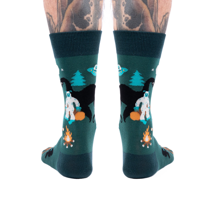 Back view of a person's lower legs with tattoos, wearing dark green cryptid socks with Yeti, Mothman, and UFO.