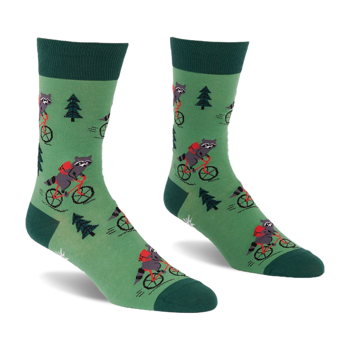 Pair of green socks with raccoons on red bicycles, wearing red backpacks, among dark green pine trees. Darker green cuffs, heels, toes.