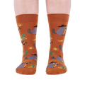 Front view of a person wearing burnt orange crew socks with a repeating pattern of raccoon cowboys, cacti, suns, and lizards.