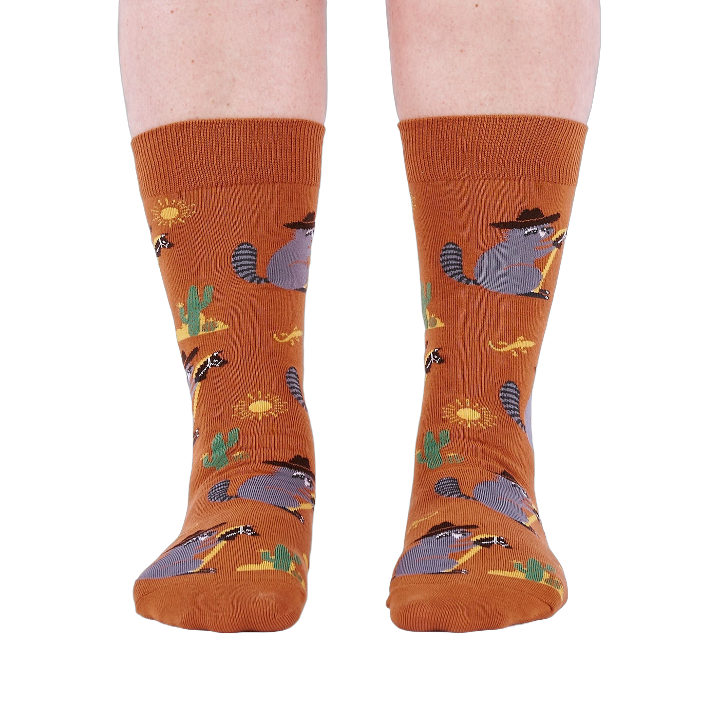 Front view of a person wearing burnt orange crew socks with a repeating pattern of raccoon cowboys, cacti, suns, and lizards.