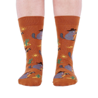 Front view of a person wearing burnt orange crew socks with a repeating pattern of raccoon cowboys, cacti, suns, and lizards.