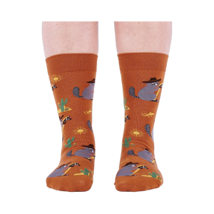 Front view of a person wearing burnt orange crew socks with a repeating pattern of raccoon cowboys, cacti, suns, and lizards.