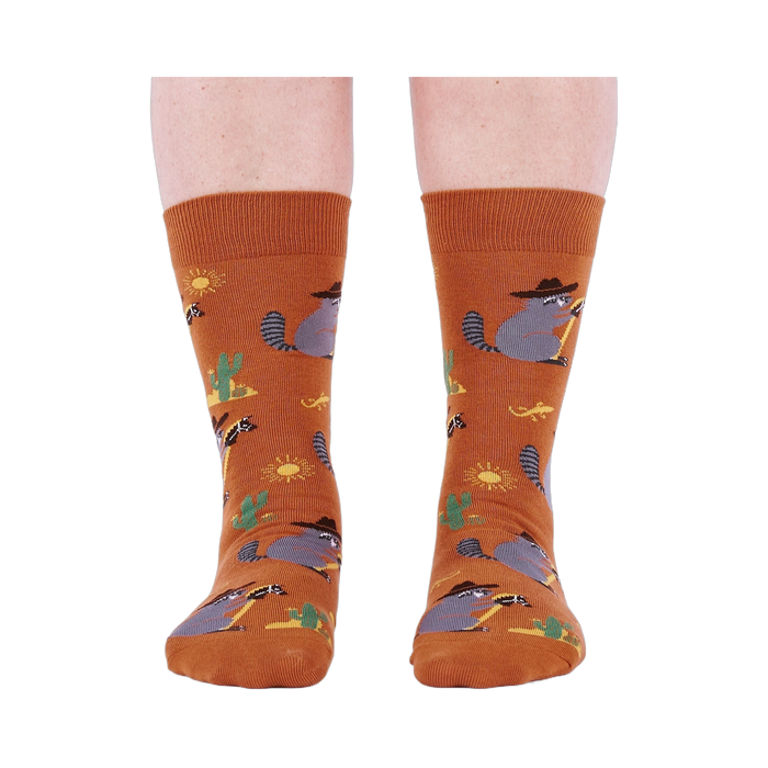 Front view of a person wearing burnt orange crew socks with a repeating pattern of raccoon cowboys, cacti, suns, and lizards.