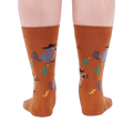 Back view of a person wearing burnt orange crew socks with a repeating pattern of raccoon cowboys, cacti, suns, and lizards.