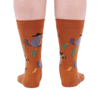 Back view of a person wearing burnt orange crew socks with a repeating pattern of raccoon cowboys, cacti, suns, and lizards.
