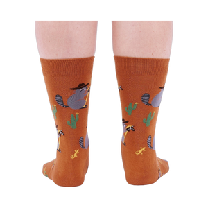 Back view of a person wearing burnt orange crew socks with a repeating pattern of raccoon cowboys, cacti, suns, and lizards.