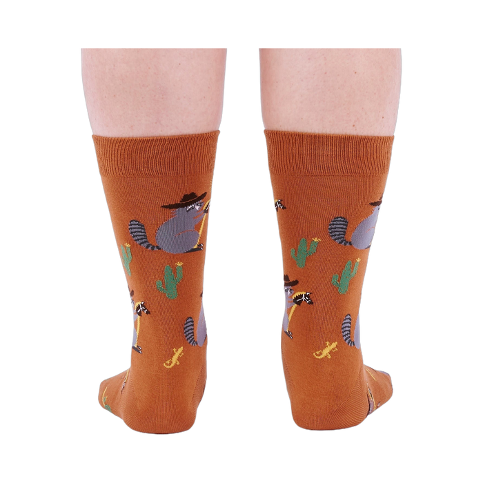 Back view of a person wearing burnt orange crew socks with a repeating pattern of raccoon cowboys, cacti, suns, and lizards.
