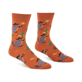 A pair of burnt orange crew socks with cartoon raccoons in cowboy hats lassoing, green cacti, yellow suns, and lizards.