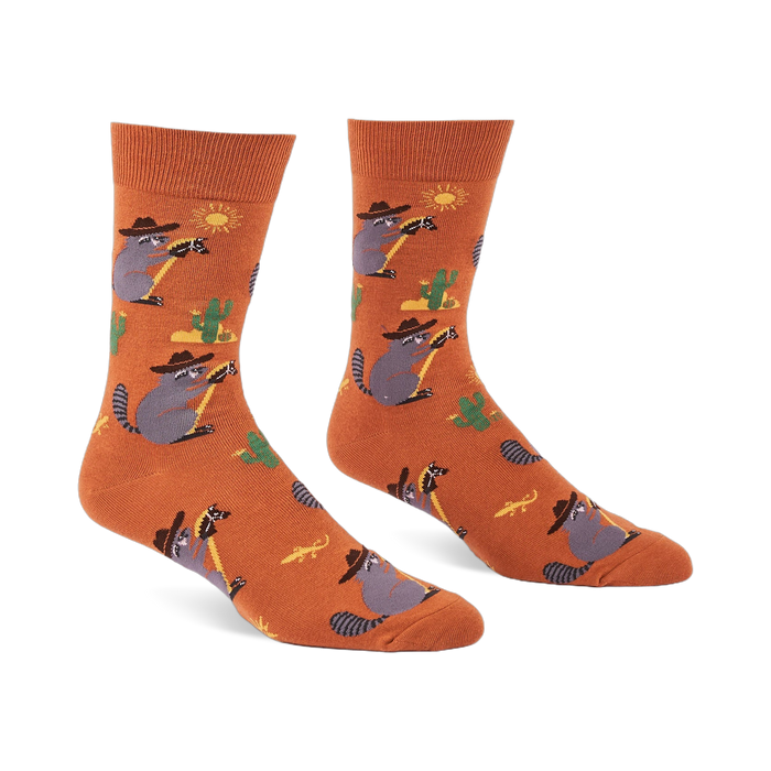 A pair of burnt orange crew socks with cartoon raccoons in cowboy hats lassoing, green cacti, yellow suns, and lizards.