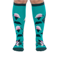 Front view of a person's legs wearing teal knee-high socks with pink and dark green philodendron leaves.