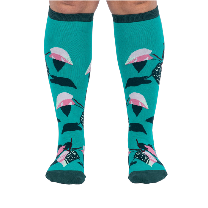 Front view of a person's legs wearing teal knee-high socks with pink and dark green philodendron leaves.