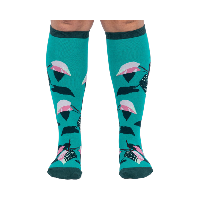 Front view of a person's legs wearing teal knee-high socks with pink and dark green philodendron leaves.