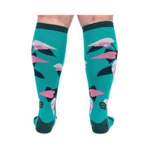 Back view of a person's legs wearing teal knee-high socks with pink and dark green philodendron leaves.