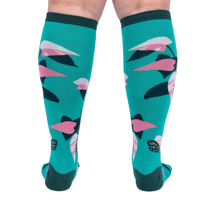 Back view of a person's legs wearing teal knee-high socks with pink and dark green philodendron leaves.