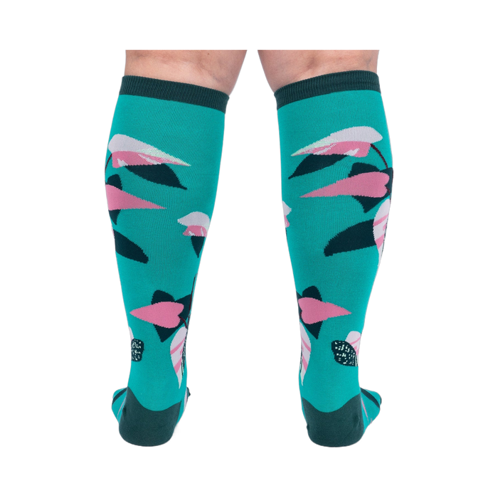 Back view of a person's legs wearing teal knee-high socks with pink and dark green philodendron leaves.