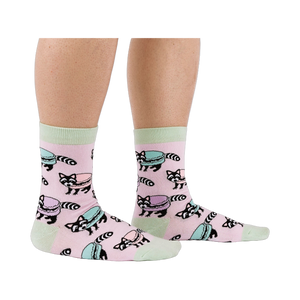 Side view of a person wearing pink crew socks with a pattern of raccoons and pastel macarons, showing light green cuffs and heels.