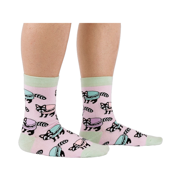 Side view of a person wearing pink crew socks with a pattern of raccoons and pastel macarons, showing light green cuffs and heels.