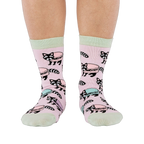 Front view of a person wearing pink crew socks adorned with a pattern of cartoon raccoons and light blue and purple macarons.