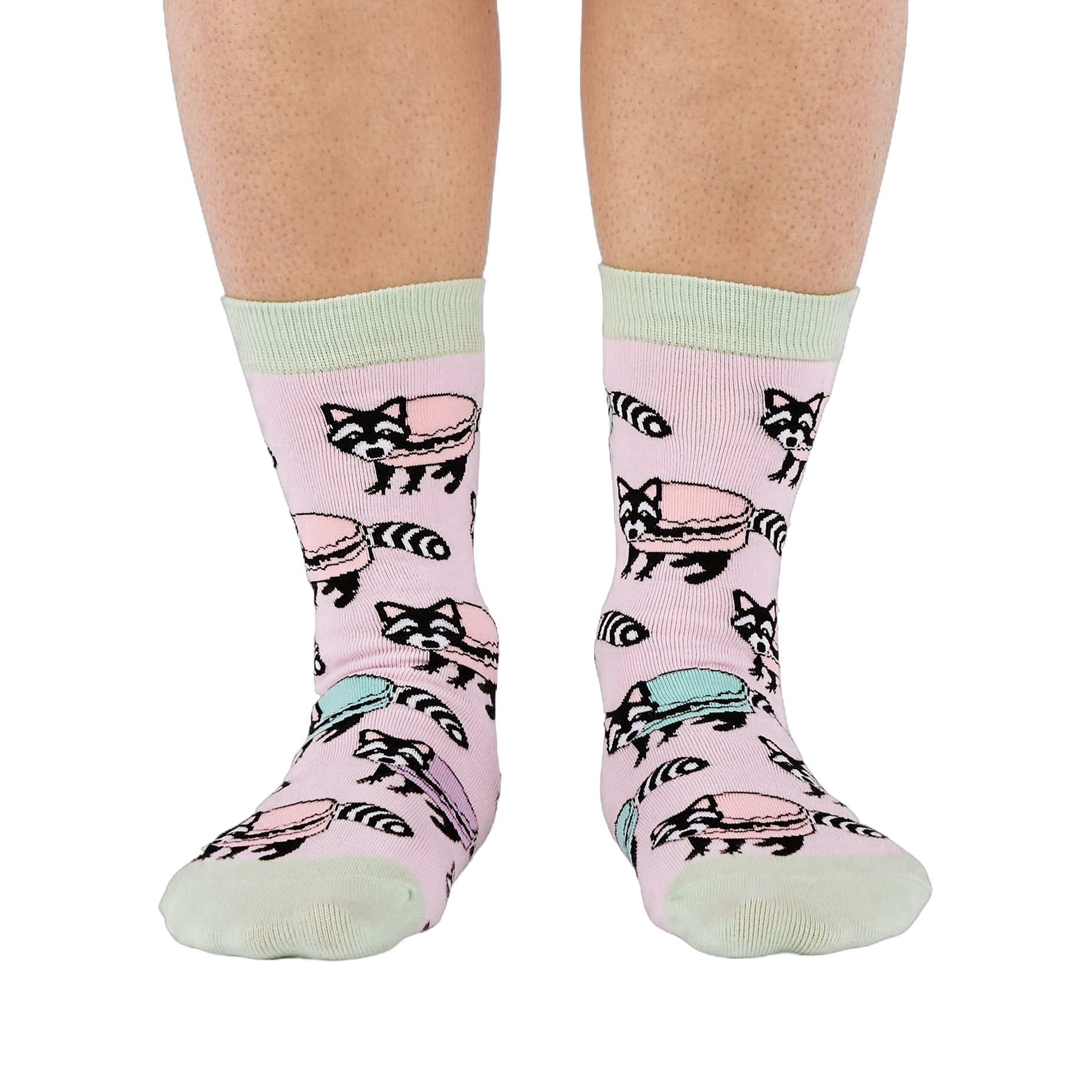 Front view of a person wearing pink crew socks adorned with a pattern of cartoon raccoons and light blue and purple macarons.