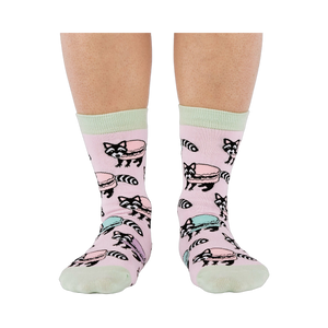Front view of a person wearing pink crew socks adorned with a pattern of cartoon raccoons and light blue and purple macarons.