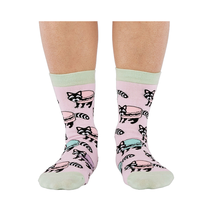 Front view of a person wearing pink crew socks adorned with a pattern of cartoon raccoons and light blue and purple macarons.