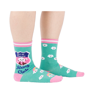 A person's legs wearing teal 'Hiking Club' crew socks, posed on a white background. Left sock shows shield design.
