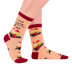 Person wearing peach "Char-cute-erie" socks, shown from the side. Socks feature smiling food characters, red hearts, and text on a light background with dark red accents.