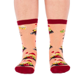 Person wearing peach "Char-cute-erie" socks, shown from the front. Socks display smiling food characters on dark boards, red hearts, and dark red cuffs, heels, and toes.