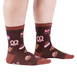 Person standing on a white floor, wearing brown crew socks patterned with cupcakes, pretzels, and chocolate.