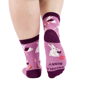 Person wearing 'Snuggle Bunny' socks, back view. Sock bottom shows 'SNUGGLE BUNNY' text and a red starburst.