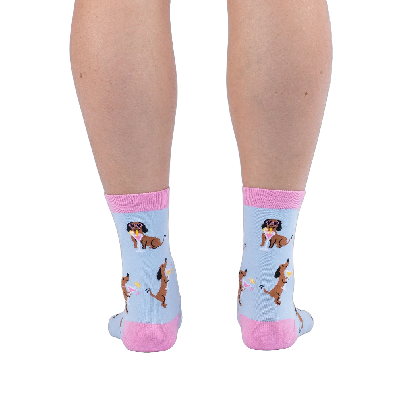 Back view of legs wearing light blue socks showing brown dachshunds wearing pink sunglasses sitting next to cocktails.