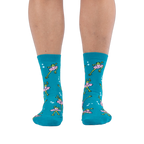 Front view of someone wearing teal crew socks featuring a pattern of green frogs in pink tutus on a black background.